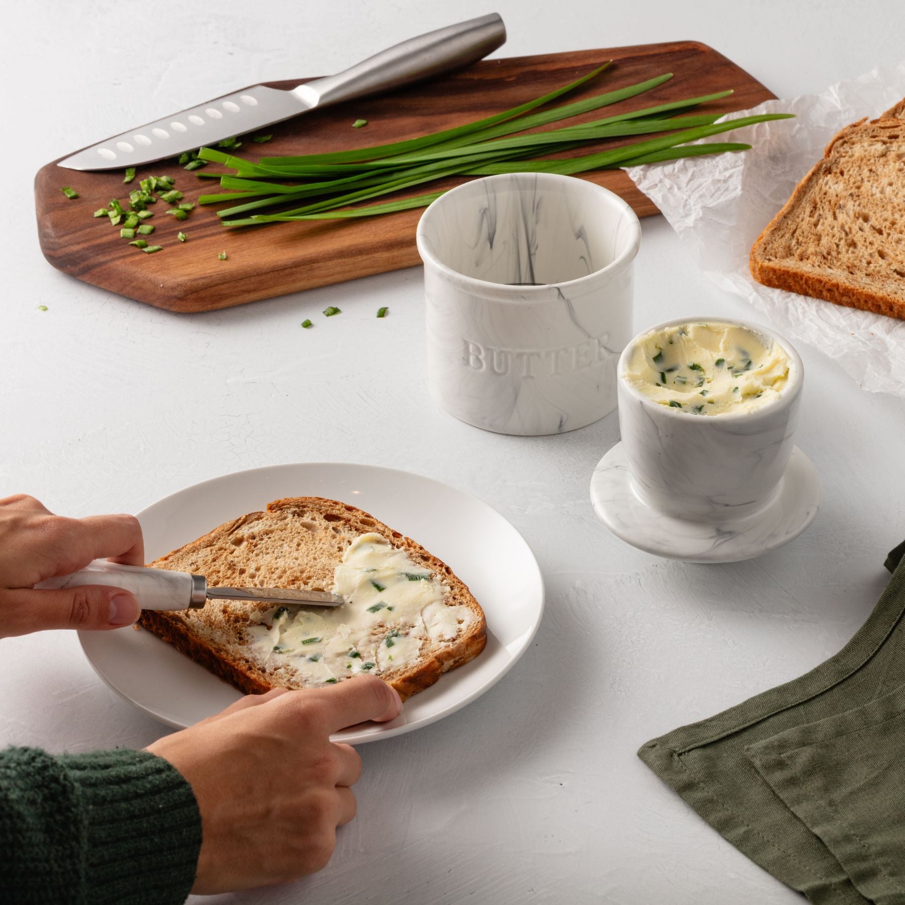 Person spreading butter on toast with a knife, surrounded by a cutting board with green onions, a marble cup, and bread.