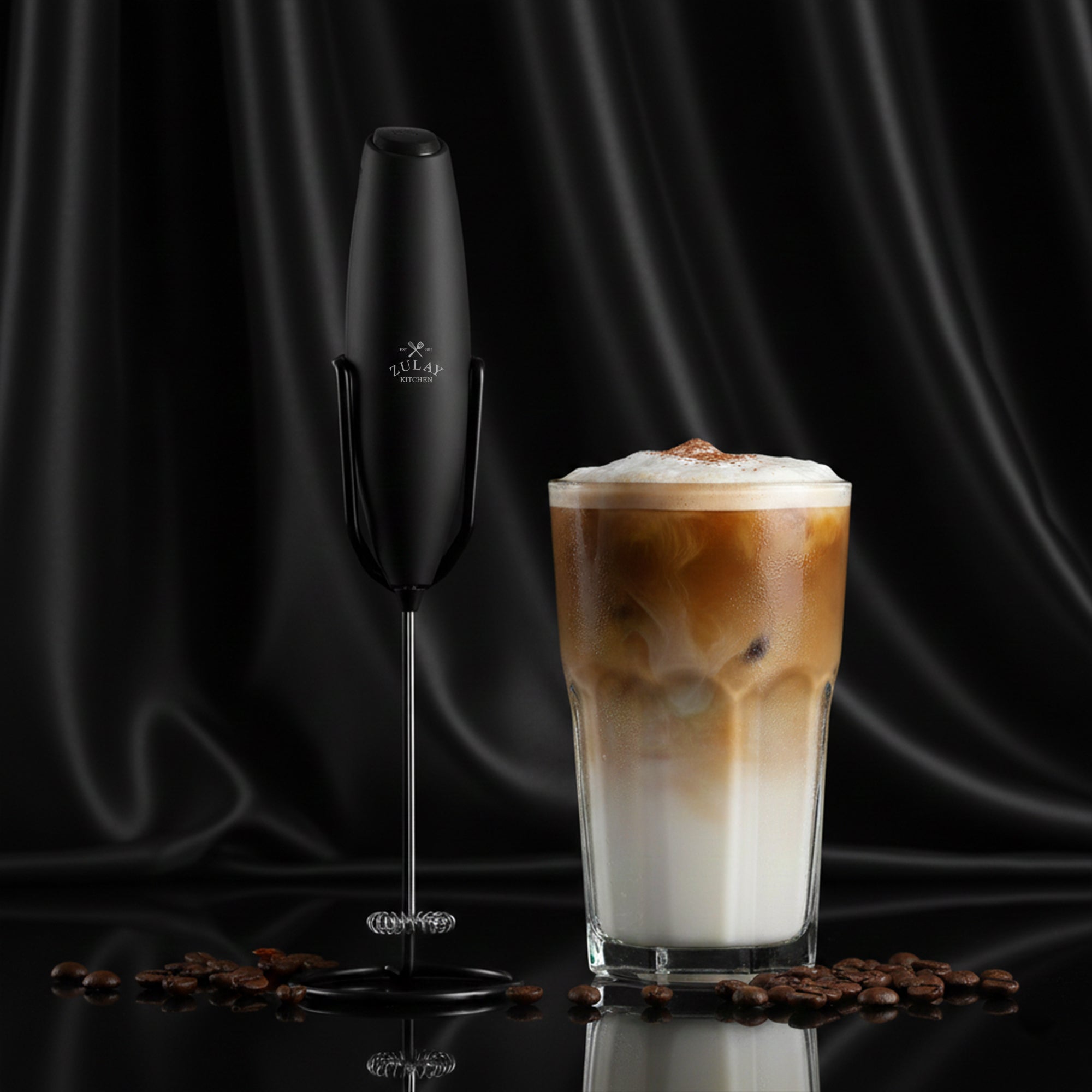 Black handheld Zulay Kitchen Executive milk frother next to a glass of iced coffee with foam on a black background