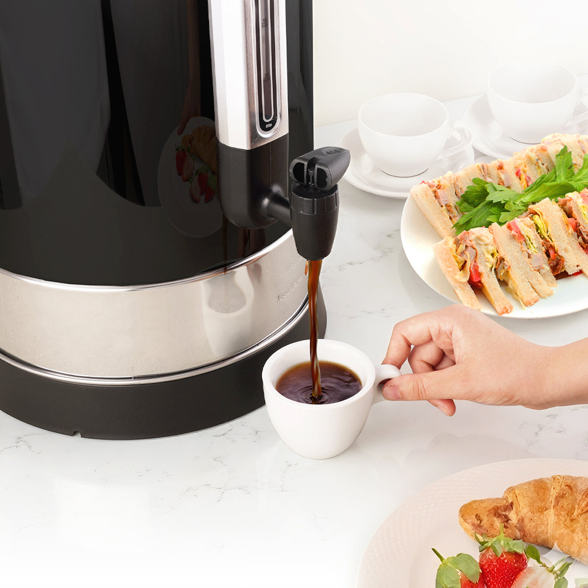 Coffee being poured from Zulay Kitchen coffee urn into a white cup on a table with sandwiches and pastries.