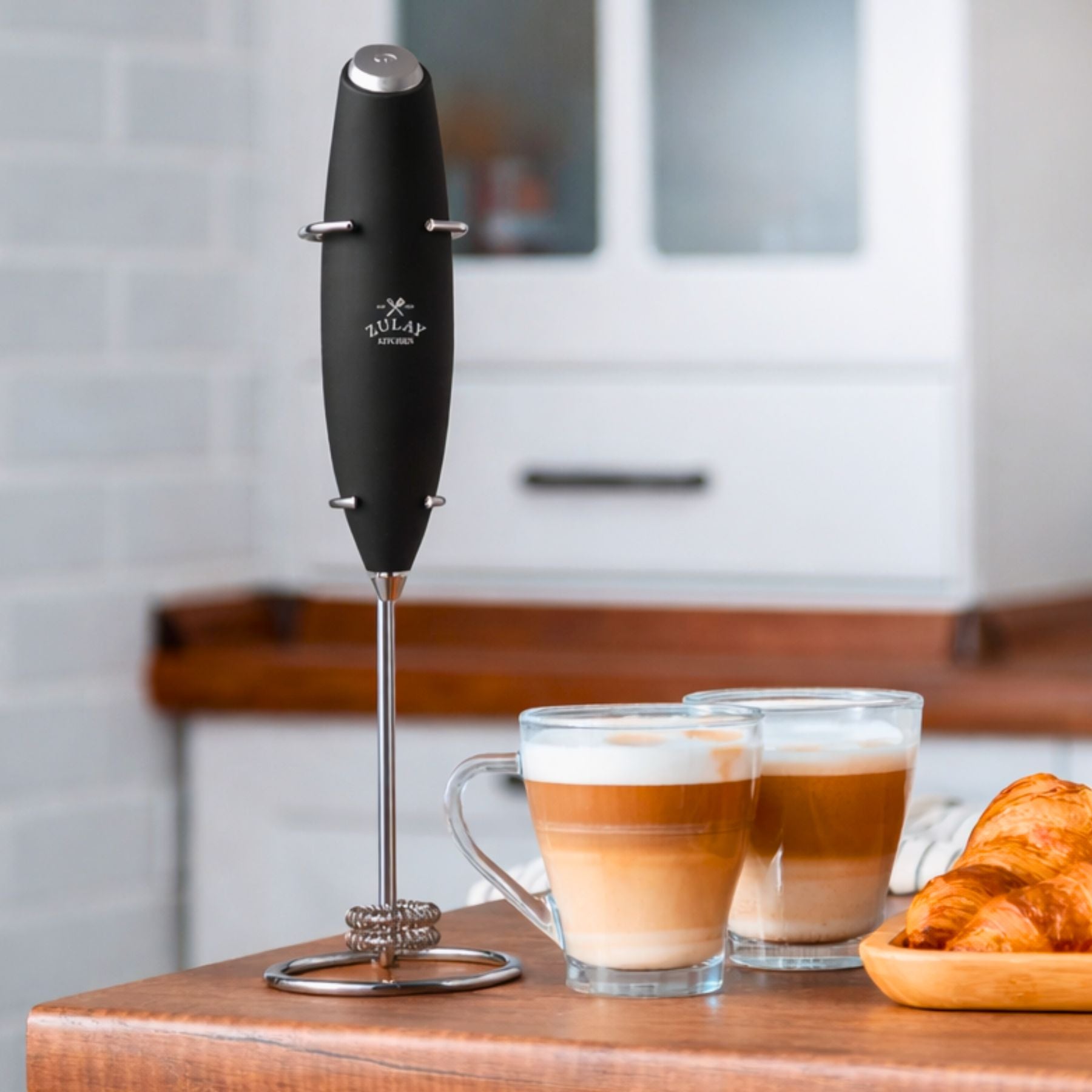Black milk frother with a stand on a kitchen counter next to two coffee cups and a croissant.