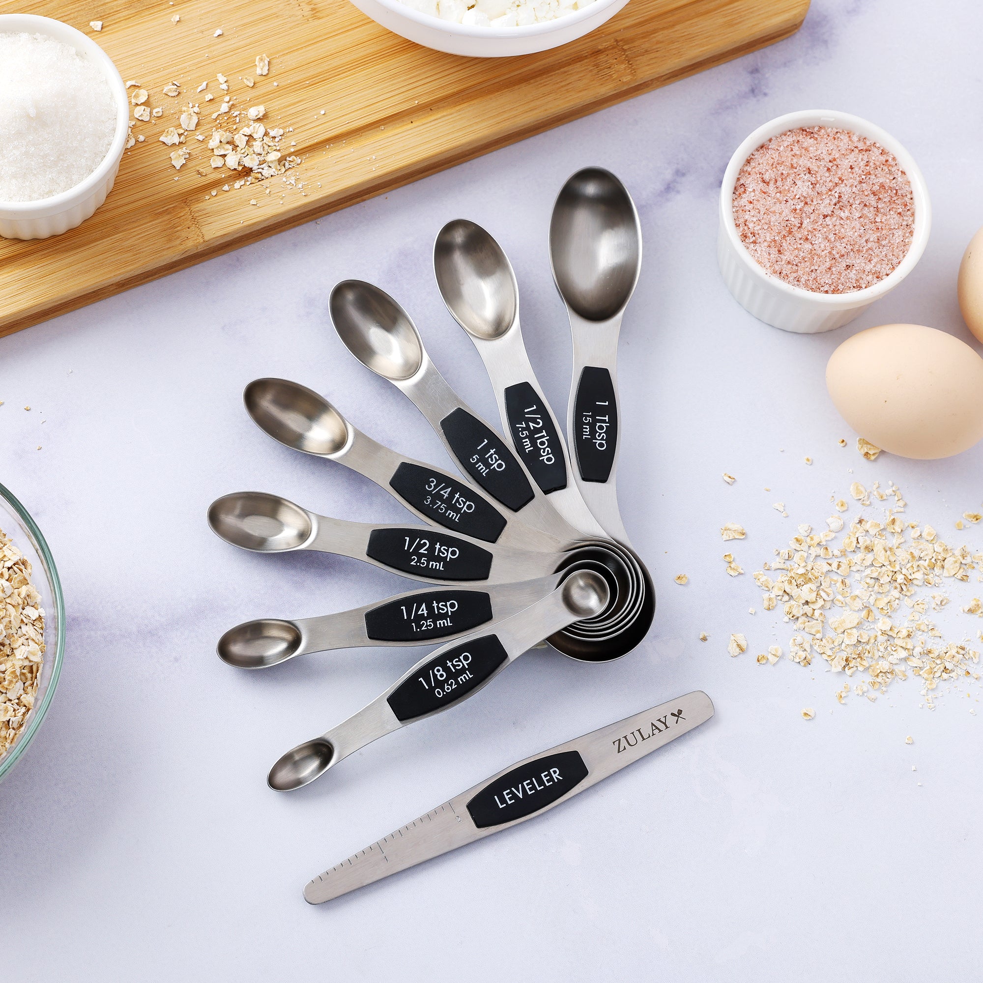 Set of stainless steel measuring spoons with black labels on a light surface with ingredients.