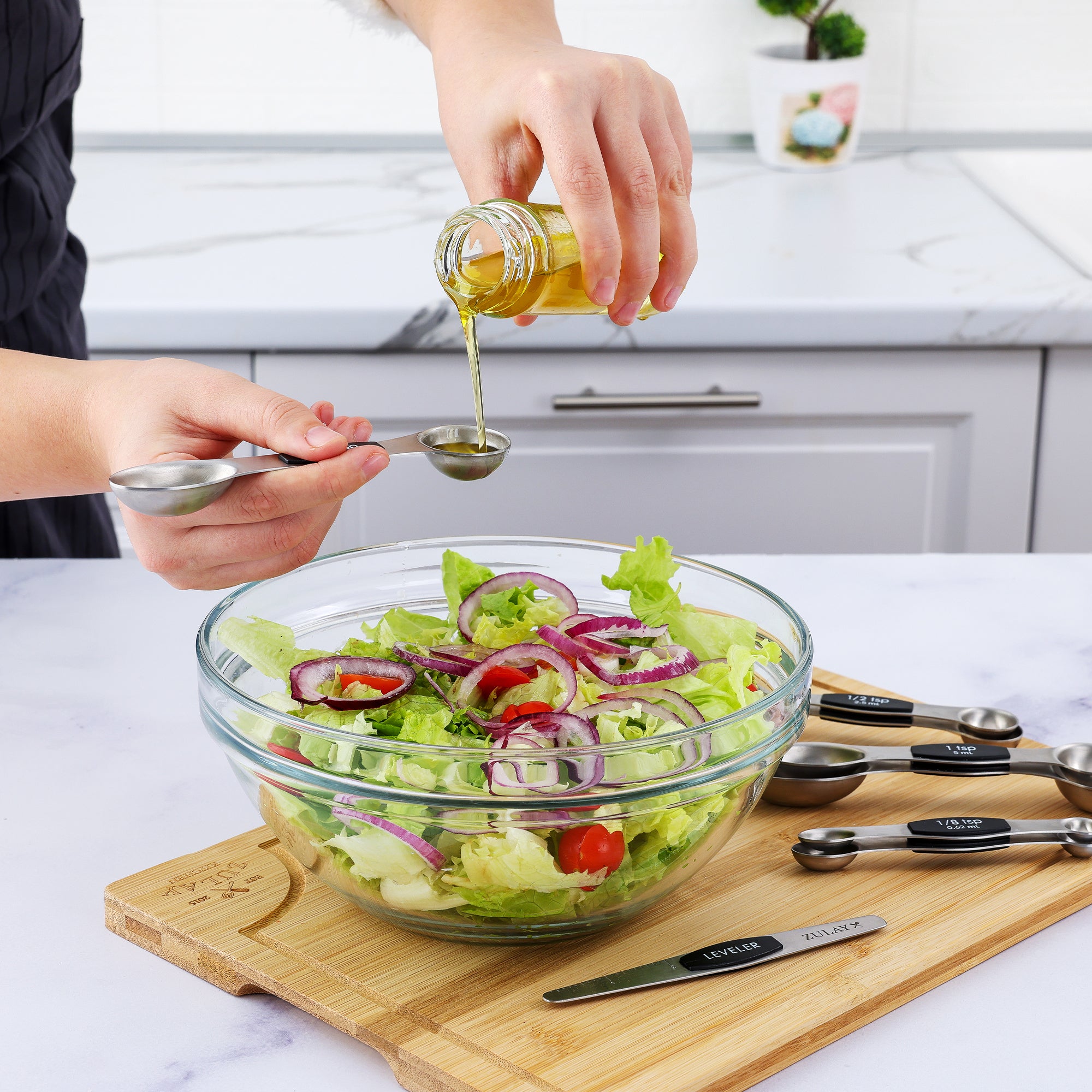 Person pouring olive oil over a salad in a glass bowl on a kitchen counter.