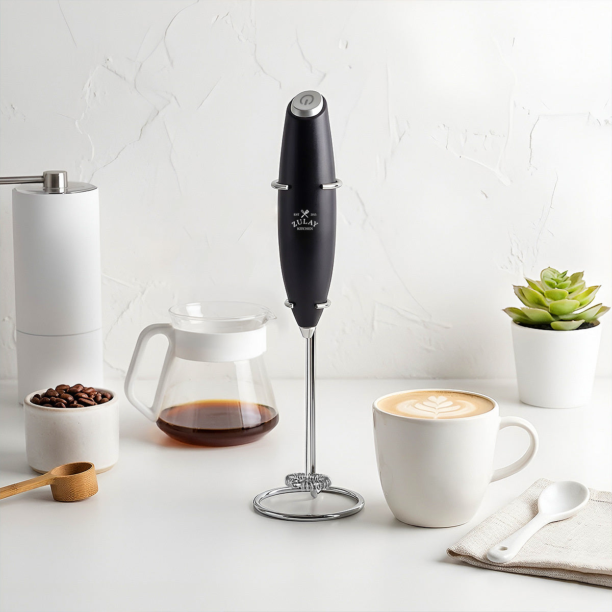 Black handheld milk frother on a kitchen counter with coffee and a plant in the background.