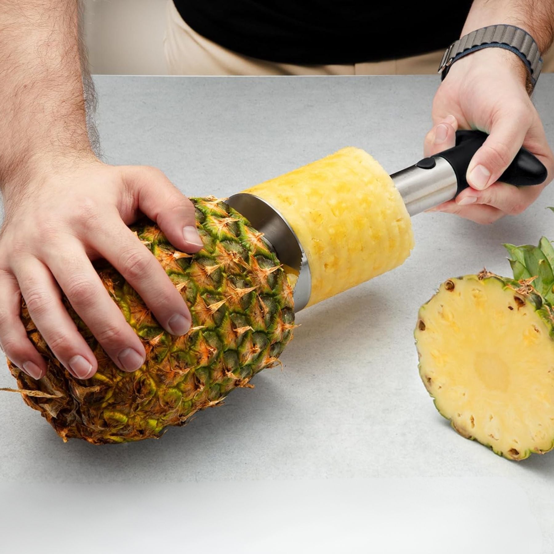 Person using a pineapple peeler to remove skin from a pineapple on a light gray surface.