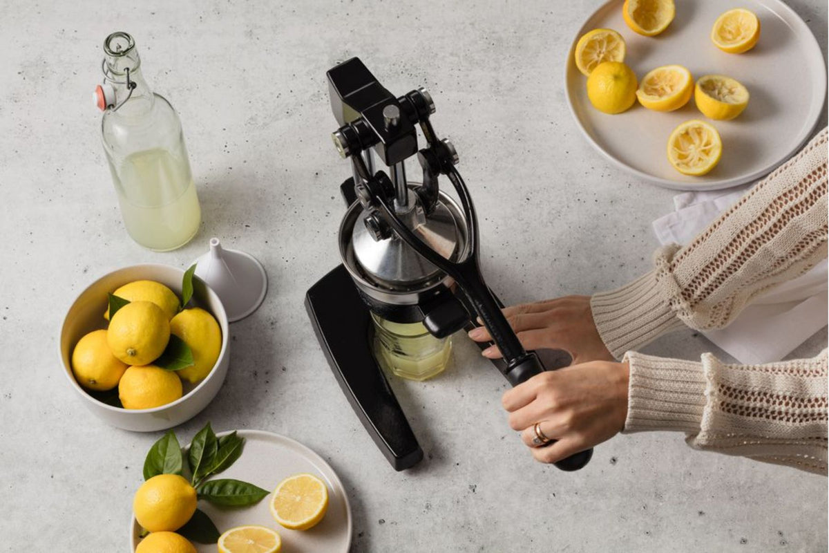 Person using a manual cast iron juicer with lemons and a glass of juice on a table.