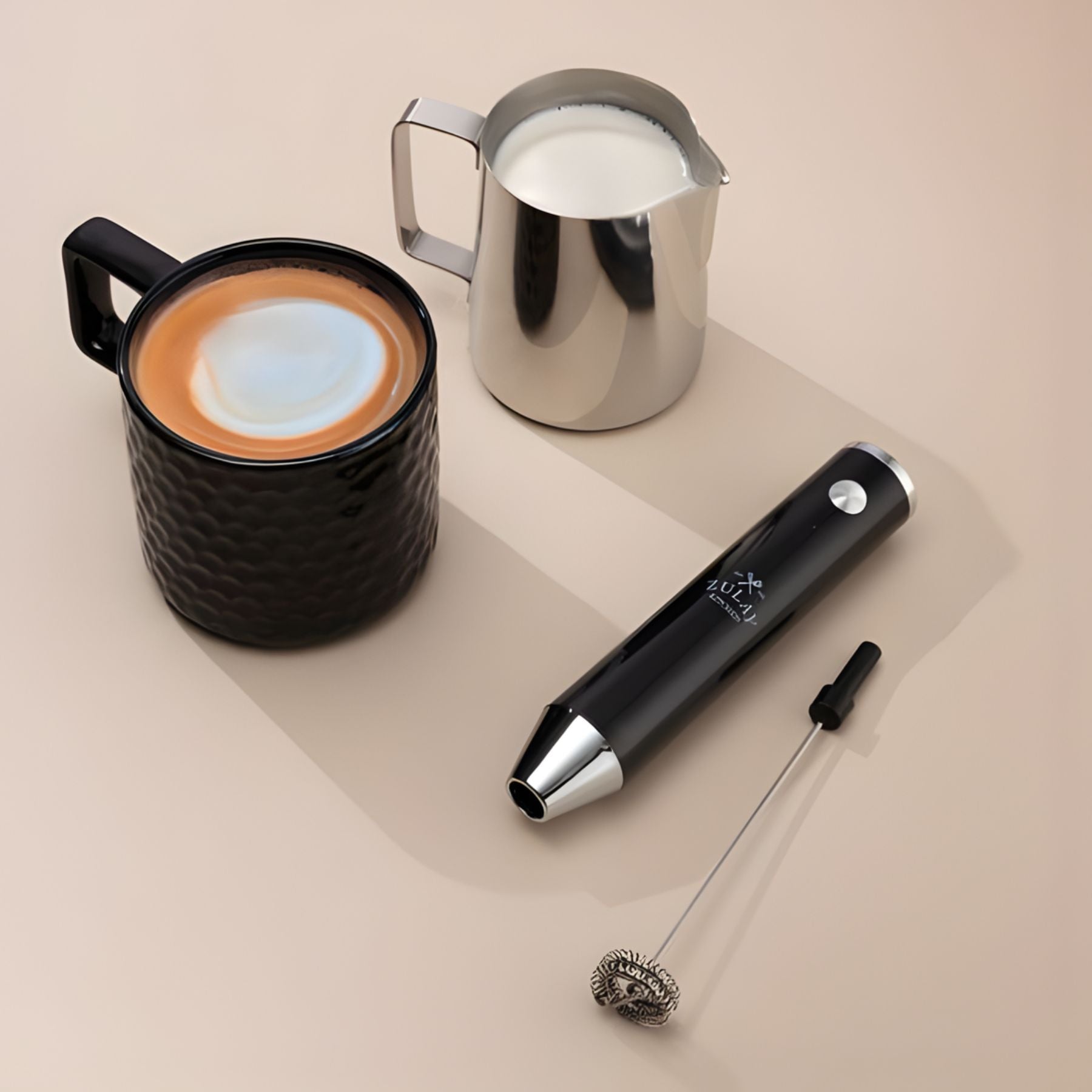 Black textured mug with a layered drink, metal milk pitcher, and black lux milk frother on a beige surface.