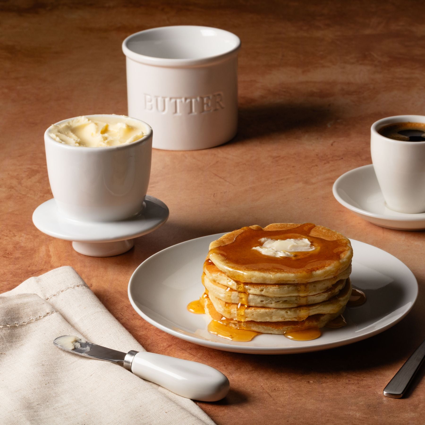 Stack of pancakes with syrup on a plate, accompanied by a cup of coffee, Zulay Kitchen butter crock set,, and knife on a wooden surface.