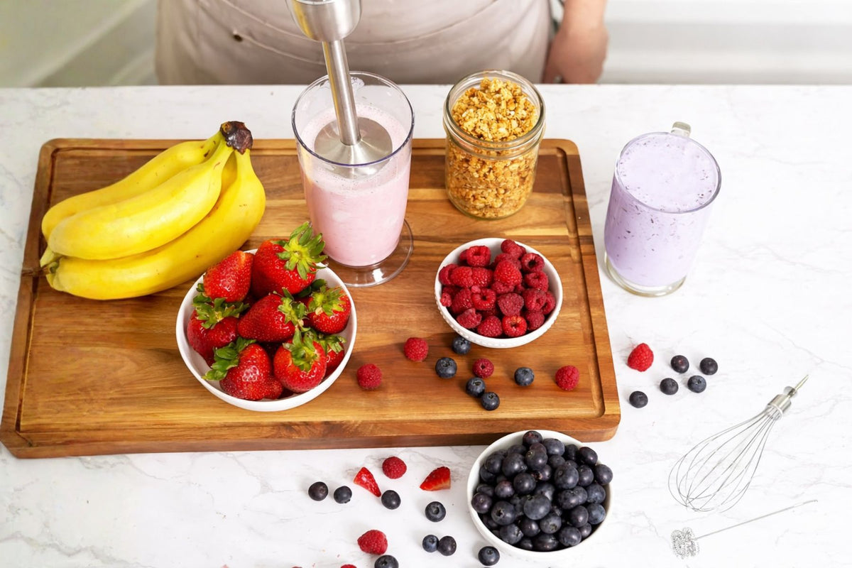 Wooden cutting board with fruits, a blender, and jars on a white surface