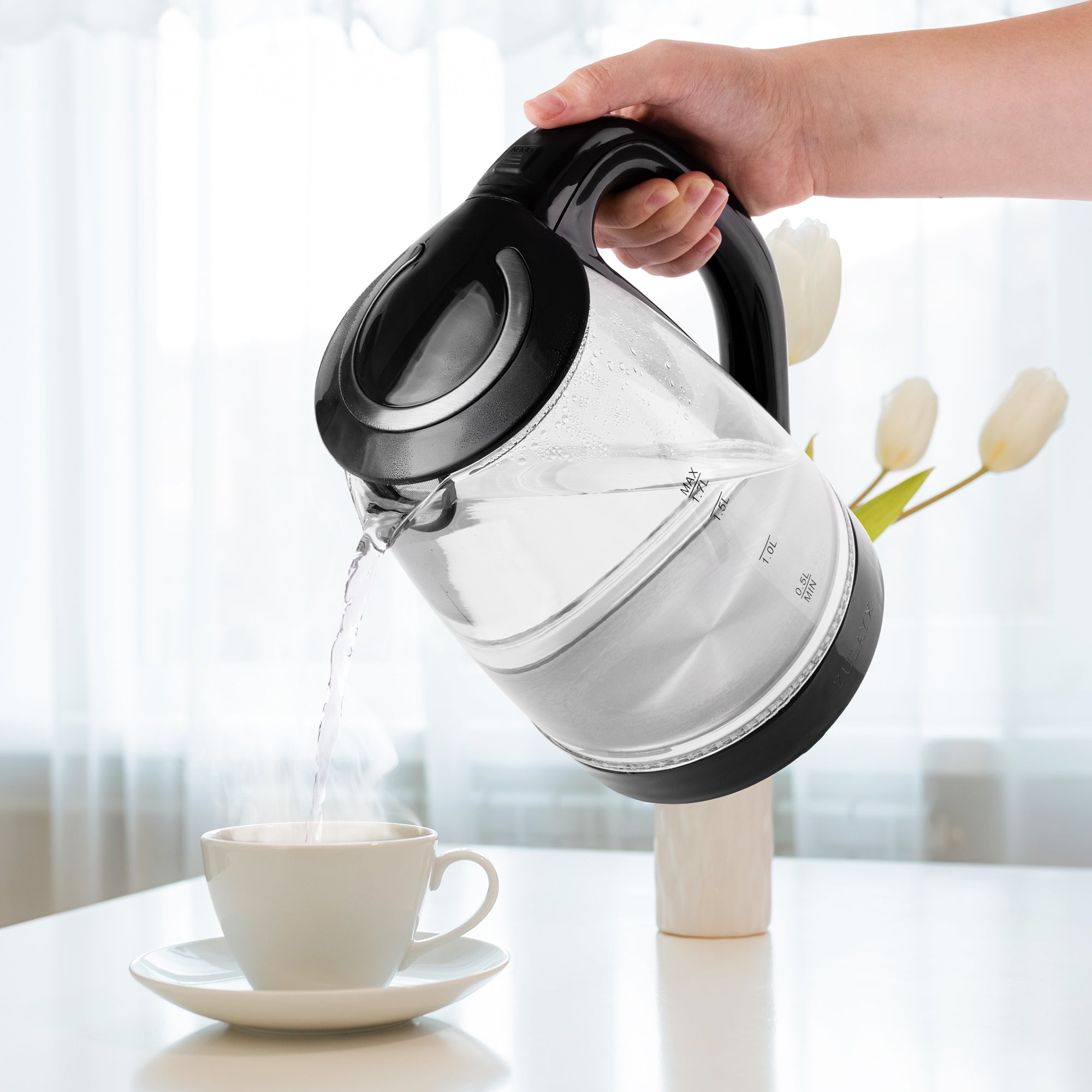 Person pouring water from the Zulay Kitchen Glass Electric kettle into a white cup on a table with a blurred background