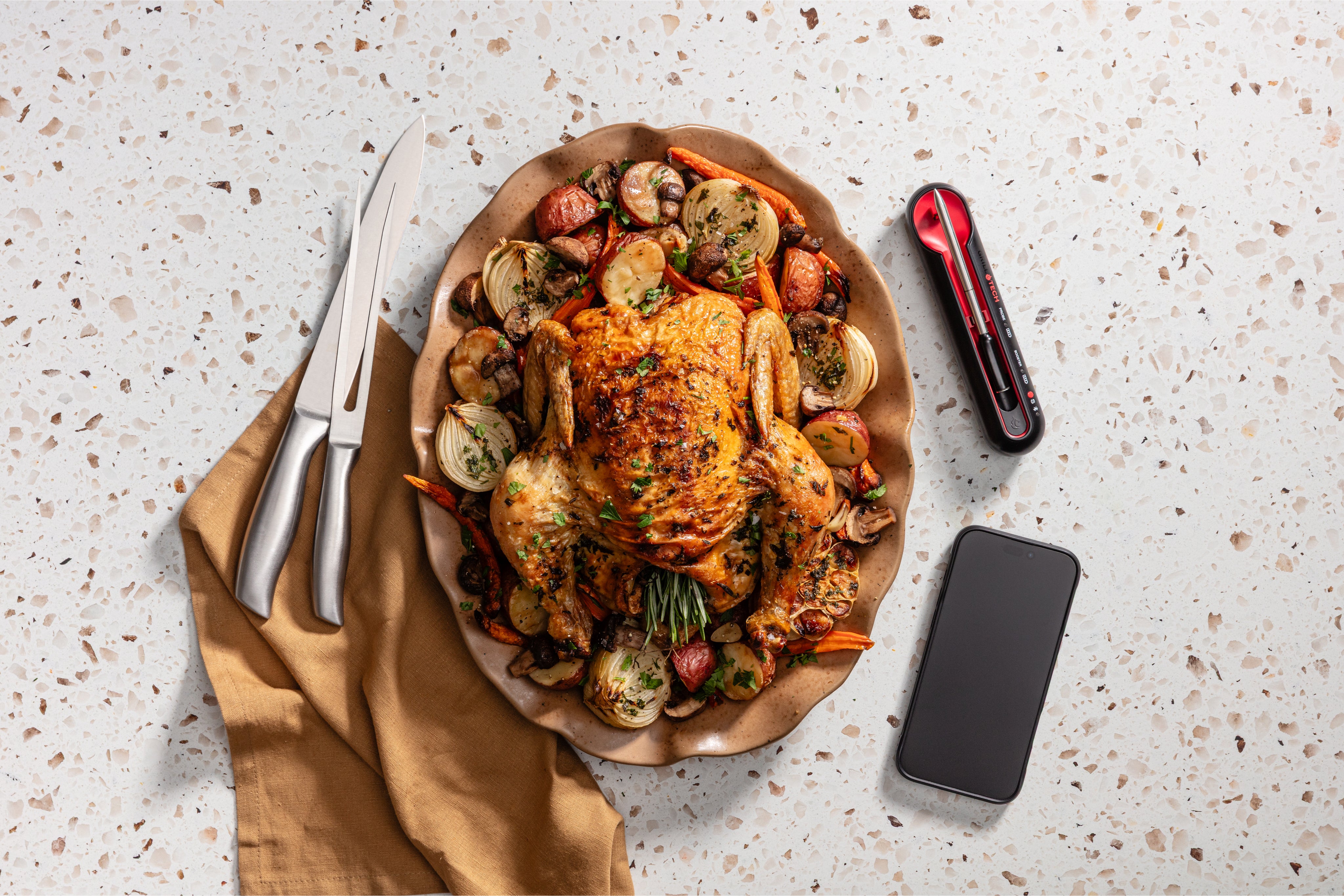 Roasted chicken with vegetables on a plate, accompanied by a smartphone and kitchen tools on a speckled countertop. With A grill tech wireless meat thermometer 