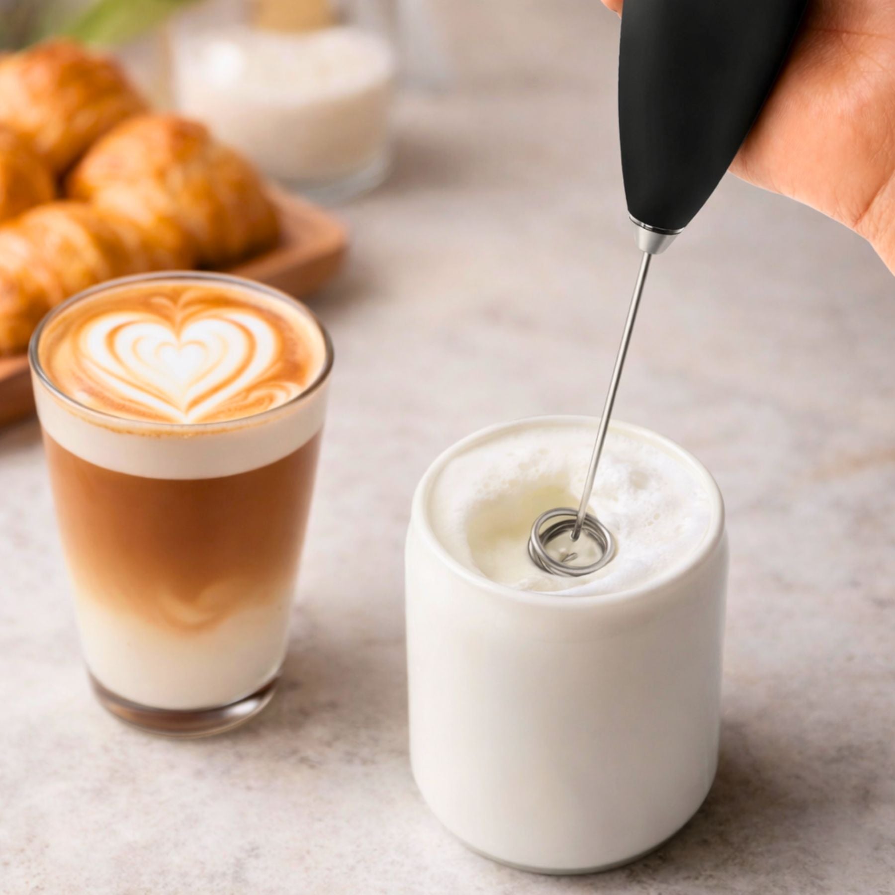 Glass of cappuccino with latte art and milk being frothed on a table.