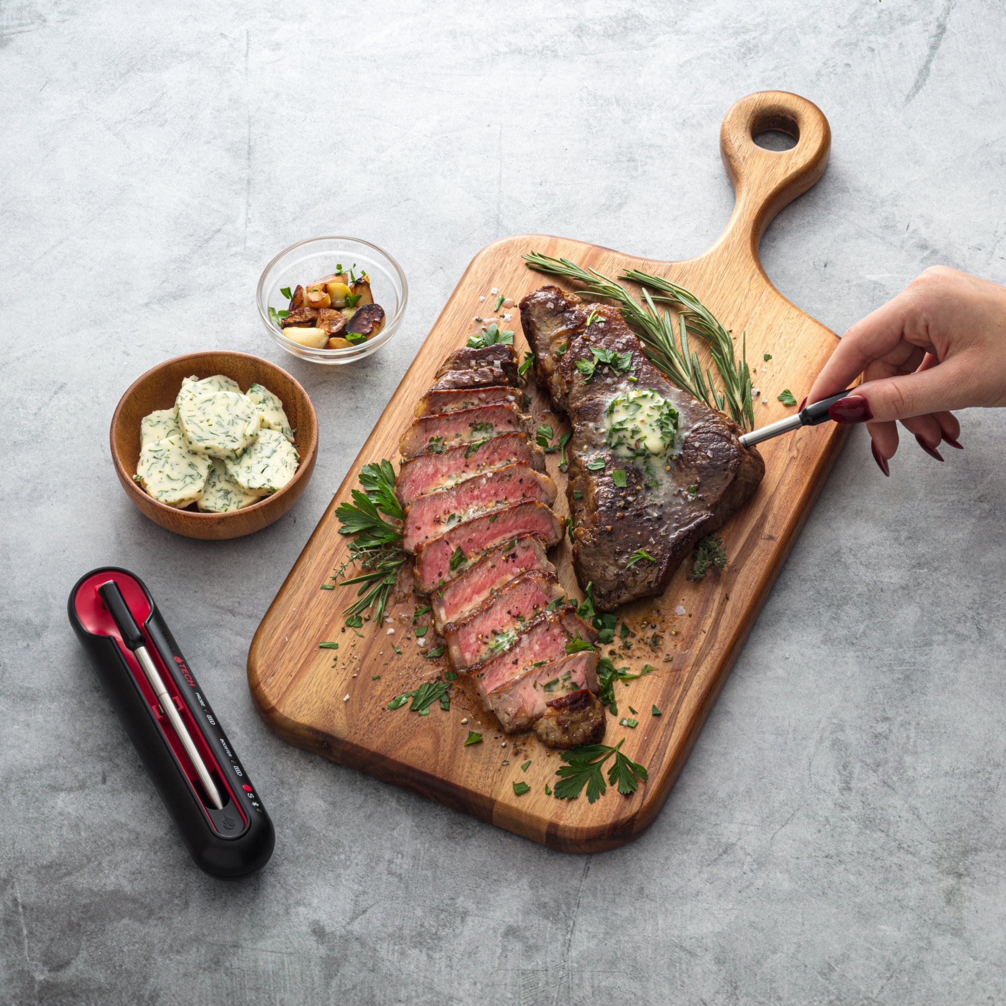 Steak on a wooden cutting board with a hand holding a knife, accompanied by a small bowl of butter and a food thermometer on a gray surface.