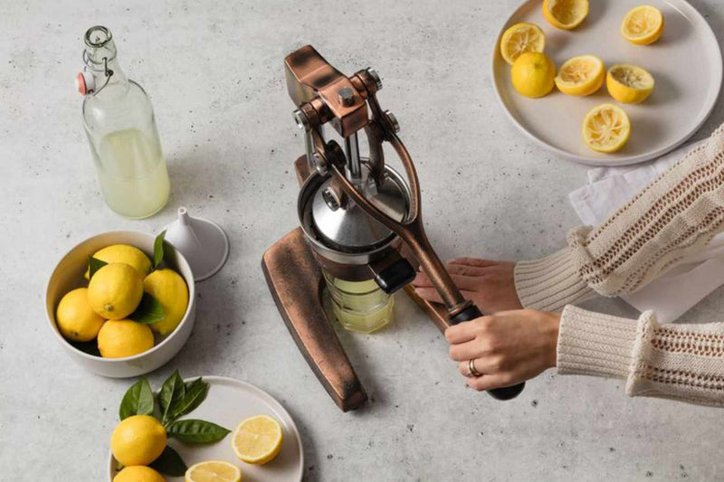 Person using a Zulay Kitchen manual lemon squeezer with lemons and a bottle of lemon juice on a table.