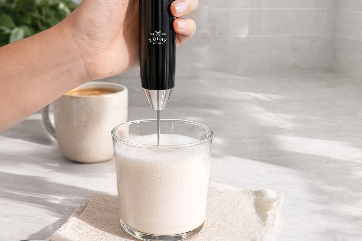 Person using a milk frother to froth milk in a glass on a kitchen counter.