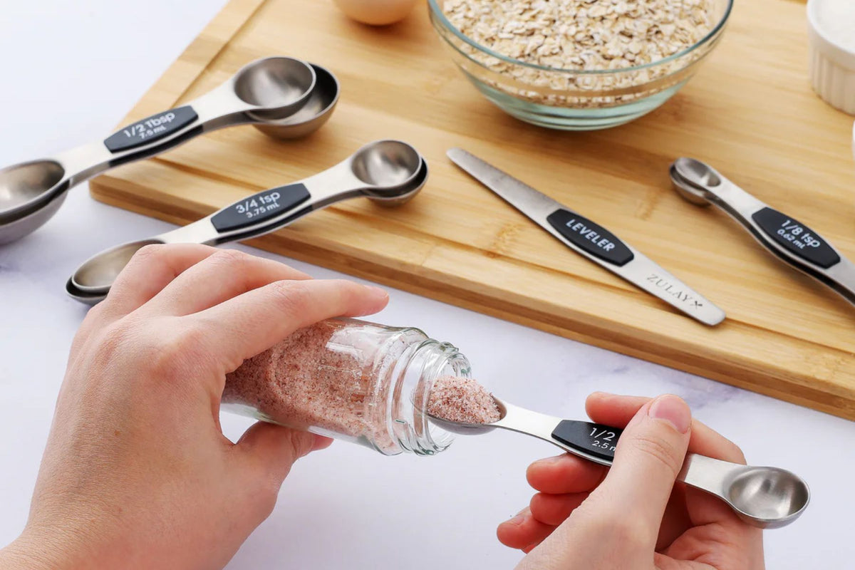 Measuring spoons being used with a wooden cutting board and ingredients in the background