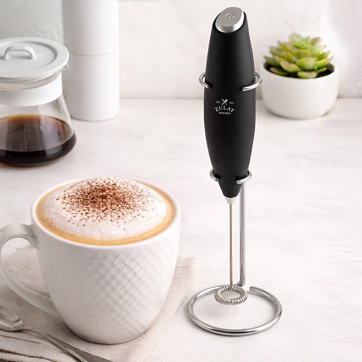 Zulay Kitchen Milk Frother With Stand next to a cup of cappuccino on a light surface.