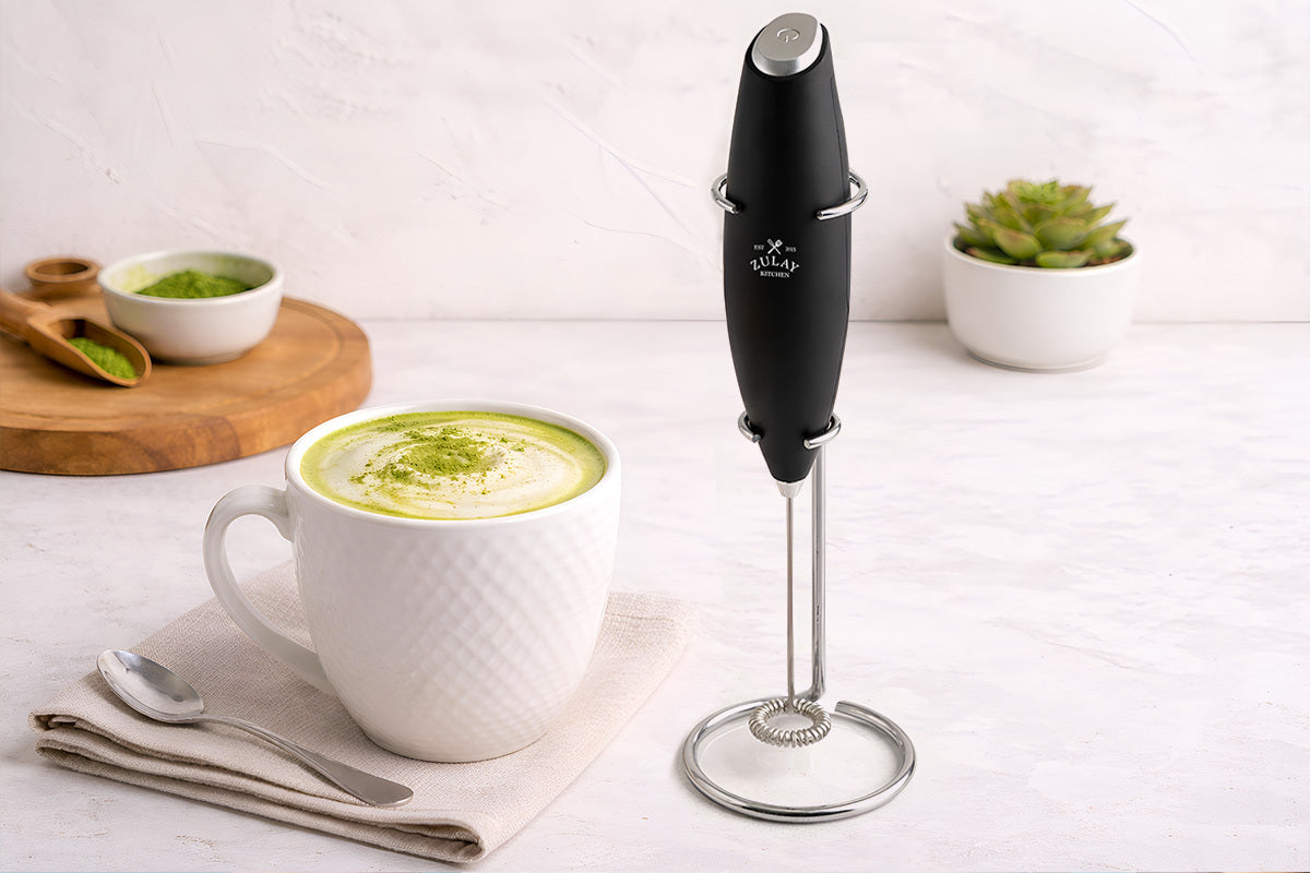 Zulay Kitchen Milk Frother With Stand next to a mug of green tea with milk on a white surface.