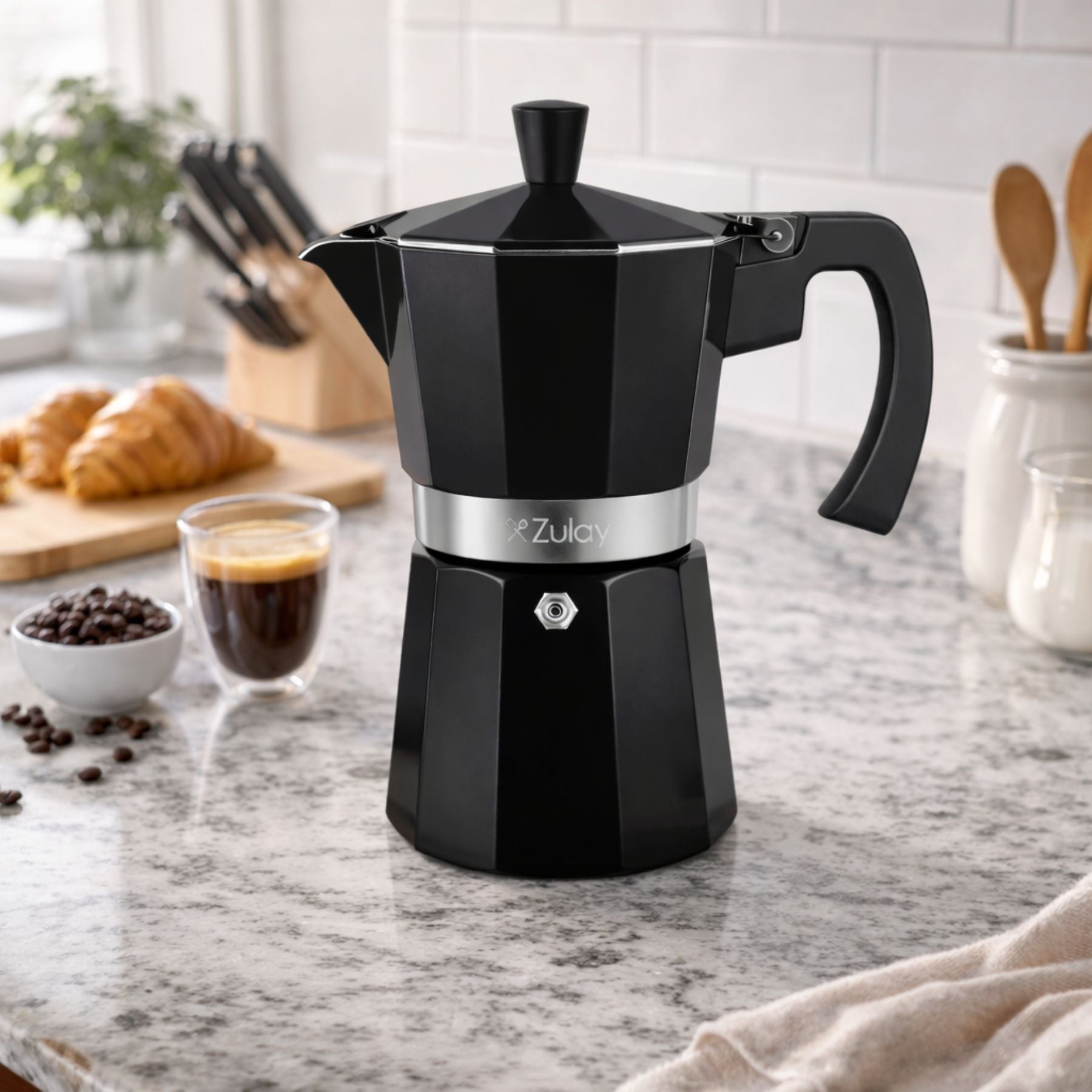 Premium Moka Pot Espresso Maker on a kitchen counter with coffee beans, a glass of coffee, and croissants in the background.