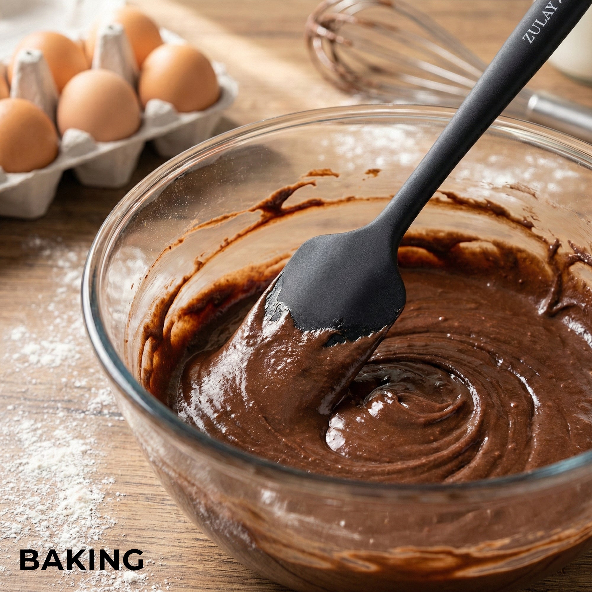 Glass bowl with chocolate batter and a Zulay Kitchen spatula, surrounded by eggs and baking tools on a wooden surface.