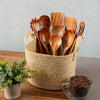 Wooden utensils in a woven basket on a wooden surface with a plant and coffee beans in the background.