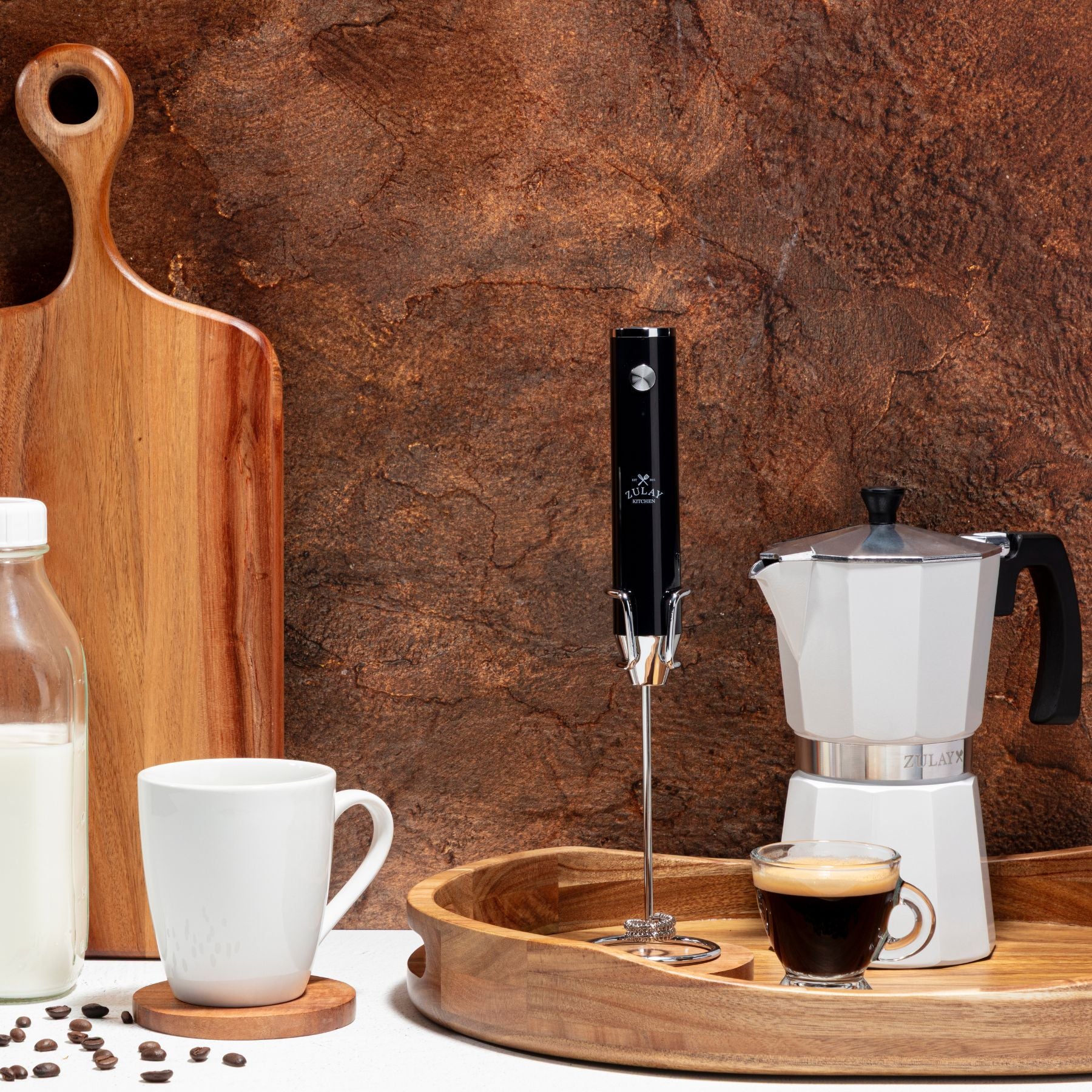 Kitchen scene with a coffee maker, milk bottle, and cup on a wooden tray against a rustic stone wall.