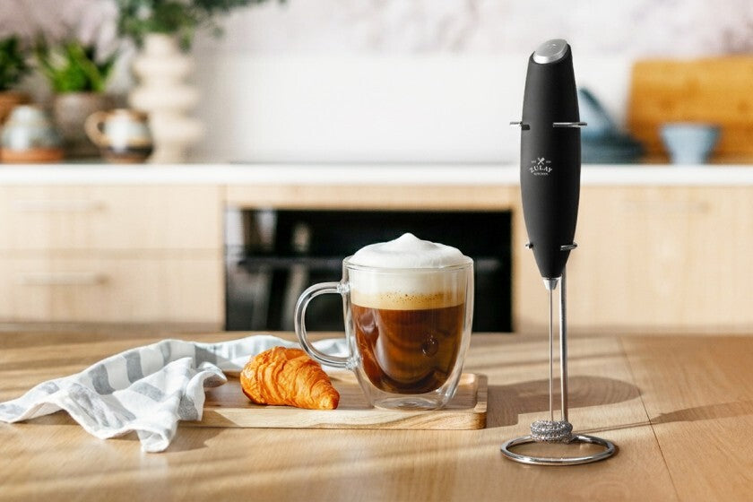 Glass of coffee with a frothy top, croissant, and Zulay Kitchen Double Whisk Milk Frother on a wooden surface.