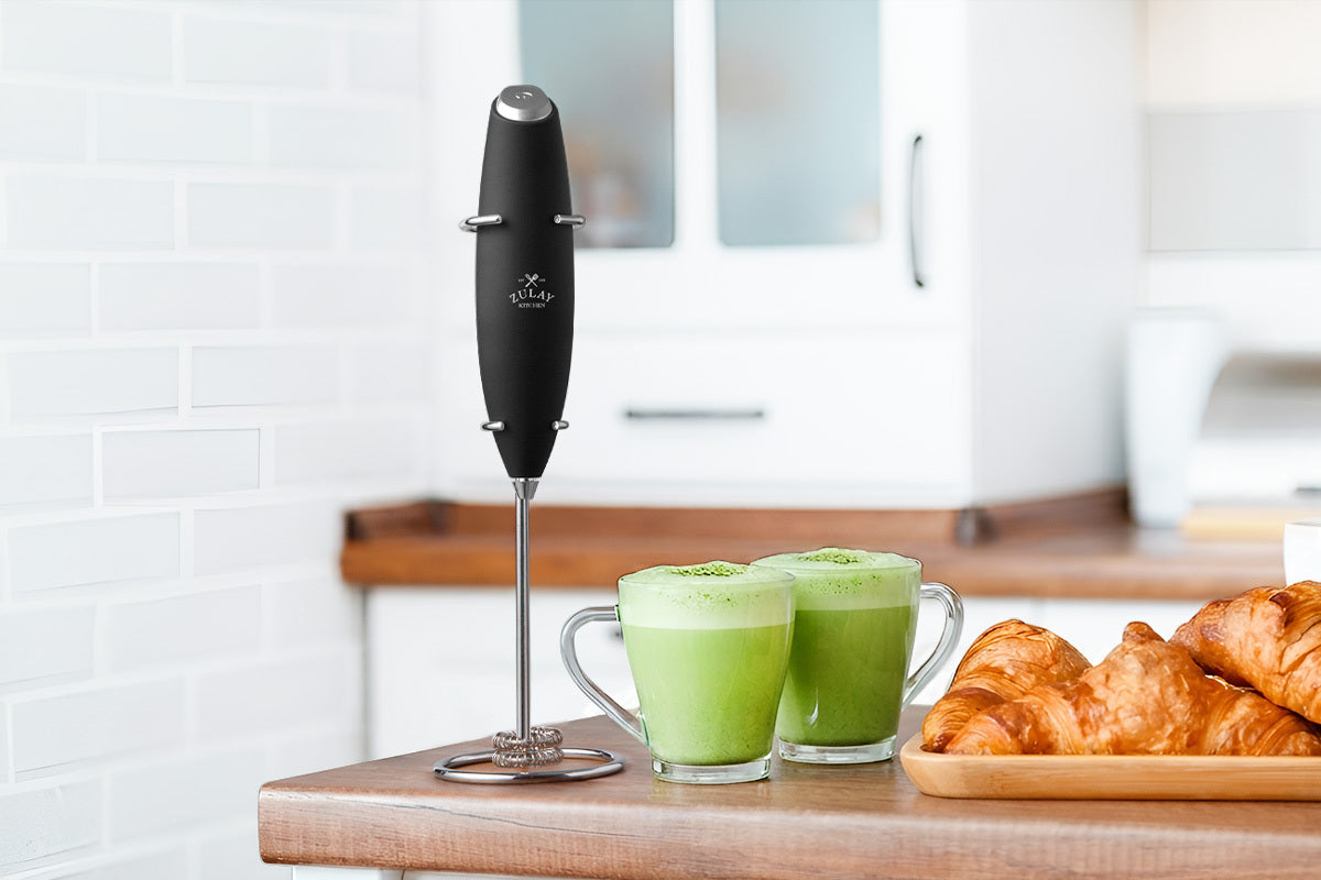 Black hand blender next to two glasses of green smoothie and a plate of croissants on a kitchen counter.