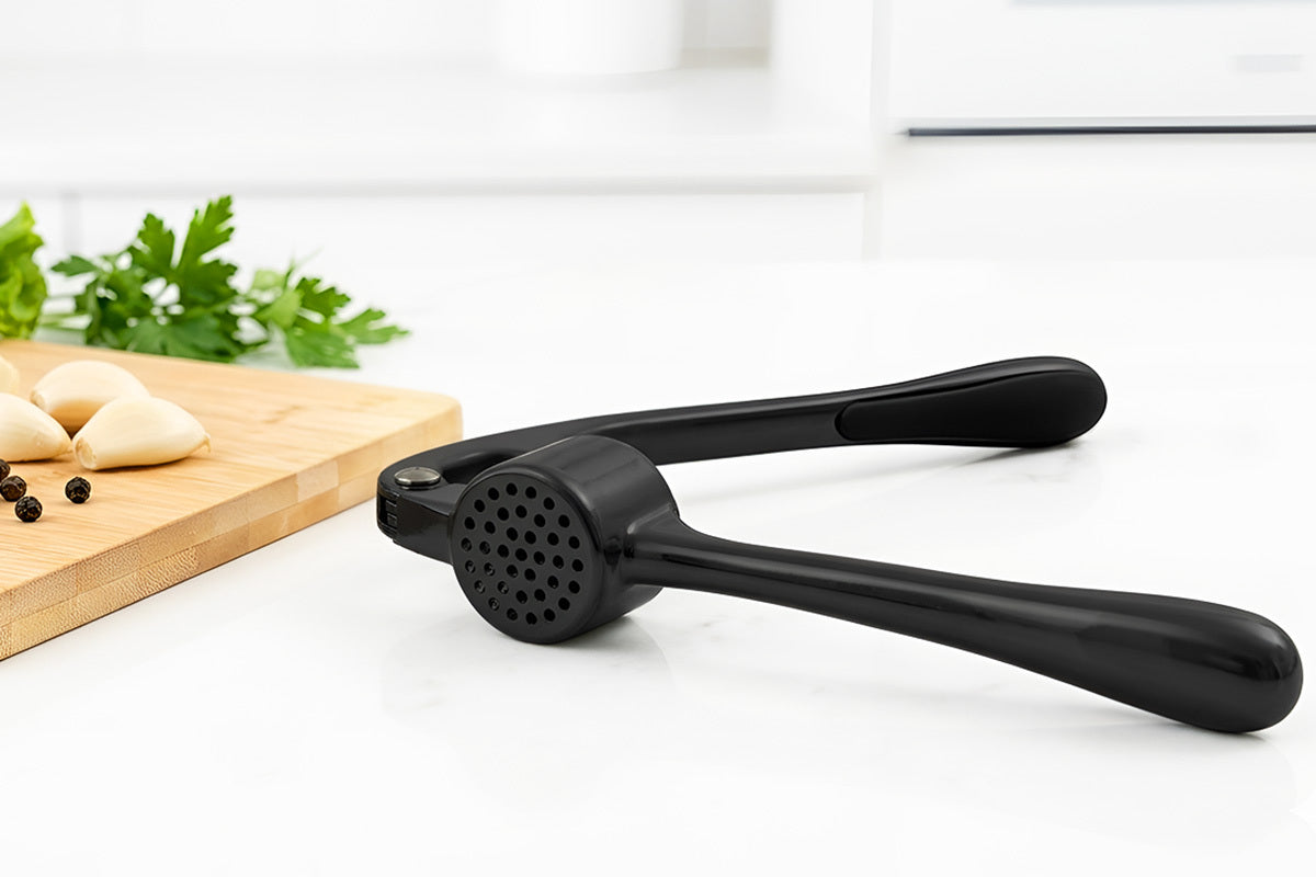 Black Zuki Garlic Press Set on a white surface with a cutting board and garlic in the background.