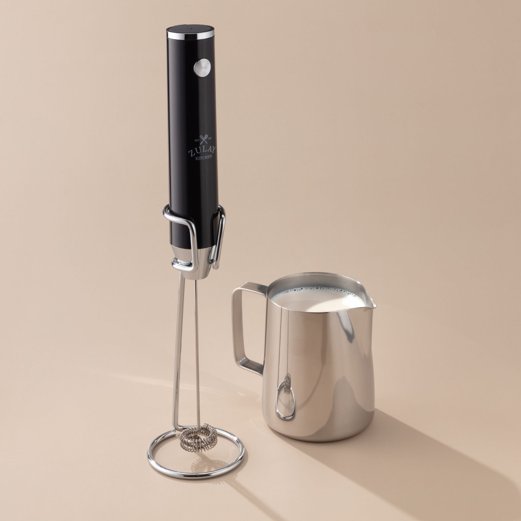 Black lux milk frother next to a stainless steel milk pitcher on a beige background